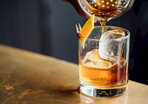 A whiskey glass with large ice cubes and an orange peel garnish being drizzled with a pour of amber liquid, on a wooden bar.