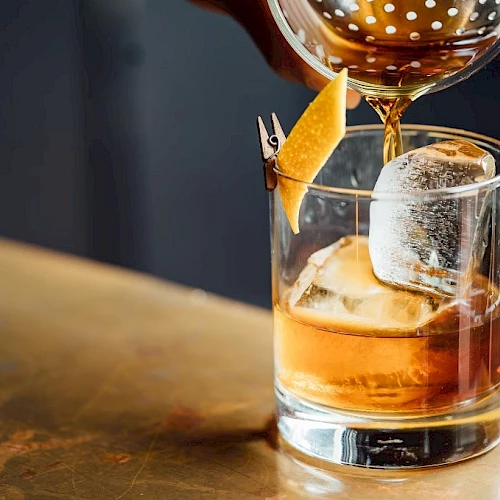 A whiskey glass with large ice cubes and an orange peel garnish being drizzled with a pour of amber liquid, on a wooden bar.