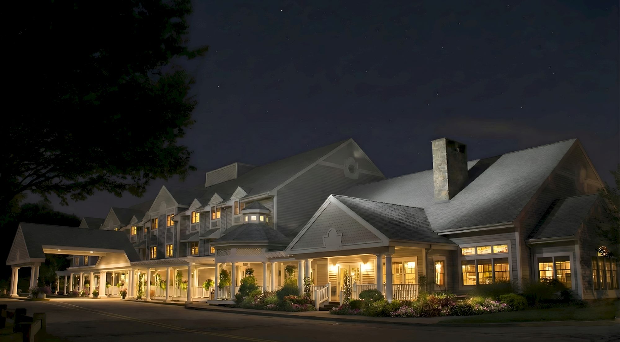 A large, cozy, well-lit suburban hotel or lodge at night with a wraparound porch, gabled roofs, and warm windows glowing.