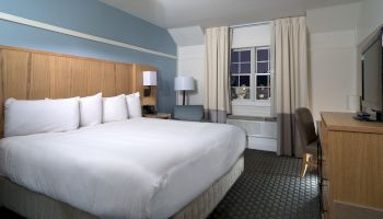 A hotel room with a large white bed, wooden headboard, nightstands, a lamp, a window with beige curtains, a desk and chair, and soft blue wall accents.
