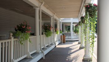 A sunlit porch with a white railing, hanging flower baskets, potted plants, and columns lining a shaded walkway; inviting and calm.