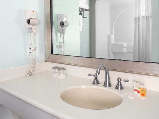 A hotel bathroom sink area with a mounted wall hair dryer, a mirror, faucet, curved sink, and small toiletries by the sink.