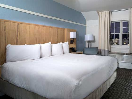 A hotel room with a large bed, white linens, wooden headboard, nightstands, a window with curtains, and a small seating/desk area.