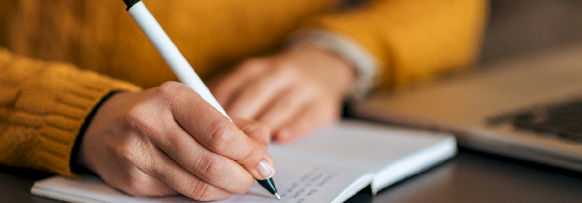 A person in a mustard sweater writes in a notebook with a pen beside a laptop, focused on jotting notes.