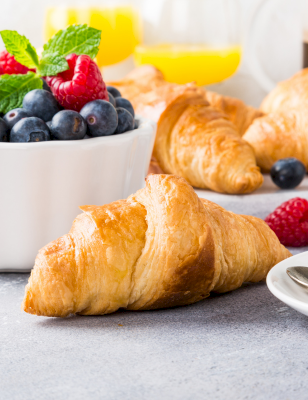 A fresh breakfast spread with croissants, berries in a bowl, coffee, orange juice, and a mint garnish on a light table.
