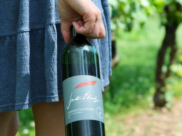 A person holding a bottle of red wine by the neck, standing outdoors in a grassy area with trees in the background.