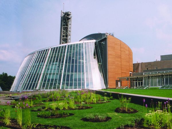 A modern glass-and-brick science/arts building with a curved glass atrium and a surrounding green garden...